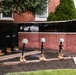 A memorial display commemorating September 11, 2001, featuring the 'Battlefield Cross' or 'Fallen Soldier Battle Cross' tribute — rifles with bayonets fixed into boots topped with helmets. The memorial is set against a brick wall engraved with names and inscribed with 'September 11, 2001' and 'We will never forget.' The display includes multiple such crosses arranged in a garden bed in front of what appears to be a public or civic building.
