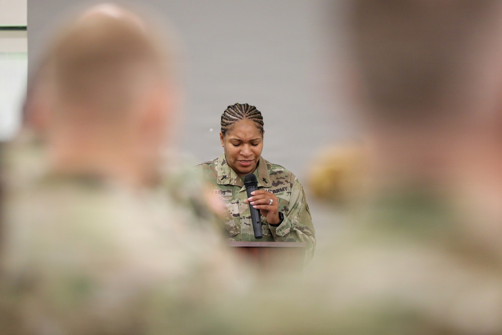 A U.S. Army soldier, a woman wearing OCP (Operational Camouflage Pattern) uniform, is speaking into a handheld microphone at what appears to be a formal military briefing or ceremony. She is photographed from behind blurred foreground figures who appear to be seated audience members in similar military uniforms. Her expression appears focused or emotional as she looks downward while speaking.
