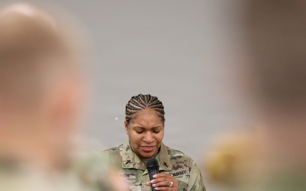 A U.S. Army soldier, a woman wearing OCP (Operational Camouflage Pattern) uniform, is speaking into a handheld microphone at what appears to be a formal military briefing or ceremony. She is photographed from behind blurred foreground figures who appear to be seated audience members in similar military uniforms. Her expression appears focused or emotional as she looks downward while speaking.