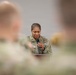 A U.S. Army soldier, a woman wearing OCP (Operational Camouflage Pattern) uniform, is speaking into a handheld microphone at what appears to be a formal military briefing or ceremony. She is photographed from behind blurred foreground figures who appear to be seated audience members in similar military uniforms. Her expression appears focused or emotional as she looks downward while speaking.