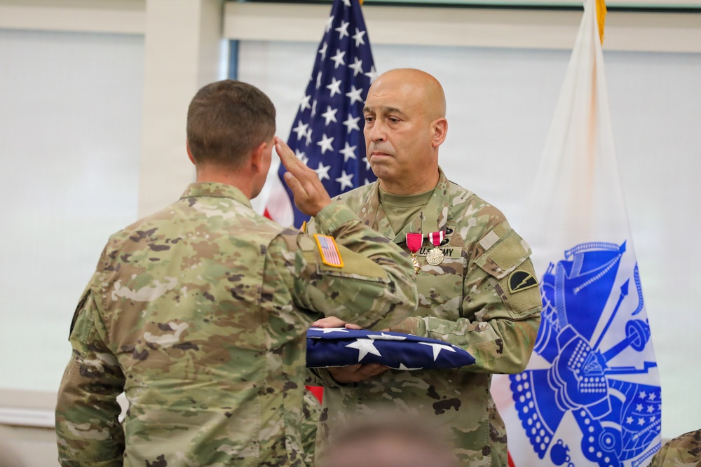 Two U.S. Army soldiers in OCP (Operational Camouflage Pattern) uniforms are engaged in a formal military ceremony. The soldier facing the camera is holding a folded American flag and saluting, while wearing medals including what appears to be a Meritorious Service Medal. The setting includes an American flag and a blue U.S. Army organizational flag in the background, suggesting a retirement or awards ceremony.