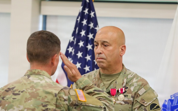 Two U.S. Army soldiers in OCP (Operational Camouflage Pattern) uniforms are engaged in a formal military ceremony. The soldier facing the camera is holding a folded American flag and saluting, while wearing medals including what appears to be a Meritorious Service Medal. The setting includes an American flag and a blue U.S. Army organizational flag in the background, suggesting a retirement or awards ceremony.