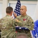 Two U.S. Army soldiers in OCP (Operational Camouflage Pattern) uniforms are engaged in a formal military ceremony. The soldier facing the camera is holding a folded American flag and saluting, while wearing medals including what appears to be a Meritorious Service Medal. The setting includes an American flag and a blue U.S. Army organizational flag in the background, suggesting a retirement or awards ceremony.
