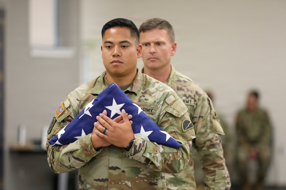 Two U.S. Army soldiers in Operational Camouflage Pattern (OCP) uniforms are participating in a formal military ceremony. The soldier in the foreground is carrying a properly folded triangular American flag against his chest with both hands, indicating a solemn occasion such as a retirement, memorial, or change of command ceremony. The expressions of both soldiers are serious and respectful, consistent with a formal military protocol.