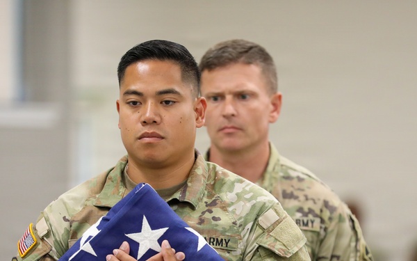 Two U.S. Army soldiers in Operational Camouflage Pattern (OCP) uniforms are participating in a formal military ceremony. The soldier in the foreground is carrying a properly folded triangular American flag against his chest with both hands, indicating a solemn occasion such as a retirement, memorial, or change of command ceremony. The expressions of both soldiers are serious and respectful, consistent with a formal military protocol.
