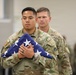 Two U.S. Army soldiers in Operational Camouflage Pattern (OCP) uniforms are participating in a formal military ceremony. The soldier in the foreground is carrying a properly folded triangular American flag against his chest with both hands, indicating a solemn occasion such as a retirement, memorial, or change of command ceremony. The expressions of both soldiers are serious and respectful, consistent with a formal military protocol.