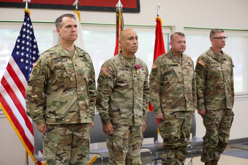 Four U.S. Army soldiers in Operational Camouflage Pattern (OCP) uniforms stand at attention during a formal military ceremony. The second soldier from the left, identified by name tape as 'FARABAUOH' (or similar), is wearing a medal on his chest, suggesting this may be an awards or change-of-command ceremony. The leftmost soldier, identified as 'SCARDELLA,' displays a brigadier general star on his chest.