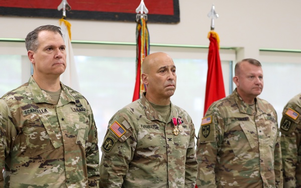 Four U.S. Army soldiers in Operational Camouflage Pattern (OCP) uniforms stand at attention during a formal military ceremony. The second soldier from the left, identified by name tape as 'FARABAUOH' (or similar), is wearing a medal on his chest, suggesting this may be an awards or change-of-command ceremony. The leftmost soldier, identified as 'SCARDELLA,' displays a brigadier general star on his chest.
