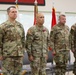 Four U.S. Army soldiers in Operational Camouflage Pattern (OCP) uniforms stand at attention during a formal military ceremony. The second soldier from the left, identified by name tape as 'FARABAUOH' (or similar), is wearing a medal on his chest, suggesting this may be an awards or change-of-command ceremony. The leftmost soldier, identified as 'SCARDELLA,' displays a brigadier general star on his chest.