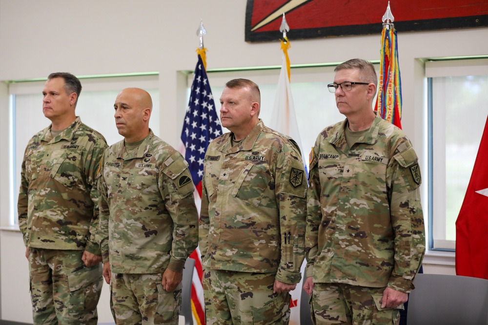 Four U.S. Army soldiers in OCP (Operational Camouflage Pattern) uniforms stand at attention in what appears to be an official military ceremony. The soldiers display various rank insignia and name tapes, with at least two appearing to hold general officer rank (visible stars). An American flag and additional military unit flags are displayed in the background.