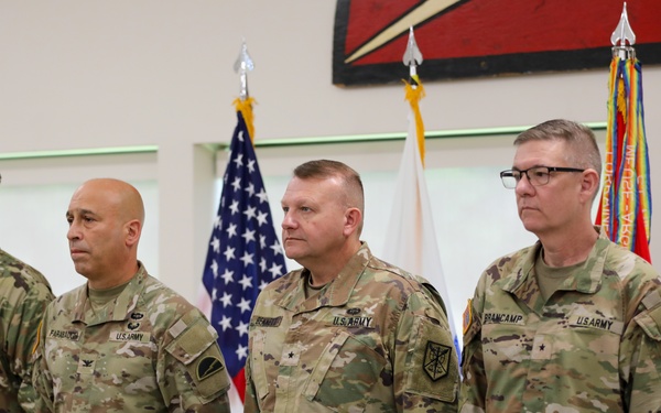 Four U.S. Army soldiers in OCP (Operational Camouflage Pattern) uniforms stand at attention in what appears to be an official military ceremony. The soldiers display various rank insignia and name tapes, with at least two appearing to hold general officer rank (visible stars). An American flag and additional military unit flags are displayed in the background.