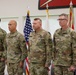 Four U.S. Army soldiers in OCP (Operational Camouflage Pattern) uniforms stand at attention in what appears to be an official military ceremony. The soldiers display various rank insignia and name tapes, with at least two appearing to hold general officer rank (visible stars). An American flag and additional military unit flags are displayed in the background.