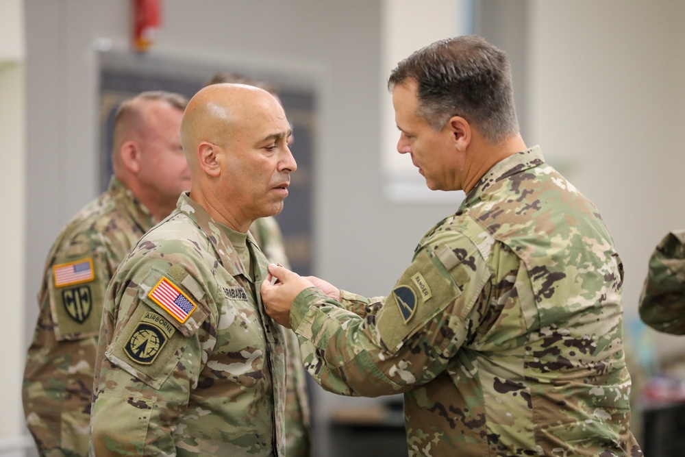 A military pinning ceremony is taking place indoors, where one U.S. Army soldier is pinning an award or insignia onto another soldier's uniform. The recipient wears Airborne and U.S. Army patches along with an OCP (Operational Camouflage Pattern) uniform. A third soldier is visible in the background observing the ceremony.