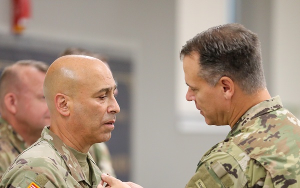 A military pinning ceremony is taking place indoors, where one U.S. Army soldier is pinning an award or insignia onto another soldier's uniform. The recipient wears Airborne and U.S. Army patches along with an OCP (Operational Camouflage Pattern) uniform. A third soldier is visible in the background observing the ceremony.