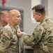 A military pinning ceremony is taking place indoors, where one U.S. Army soldier is pinning an award or insignia onto another soldier's uniform. The recipient wears Airborne and U.S. Army patches along with an OCP (Operational Camouflage Pattern) uniform. A third soldier is visible in the background observing the ceremony.