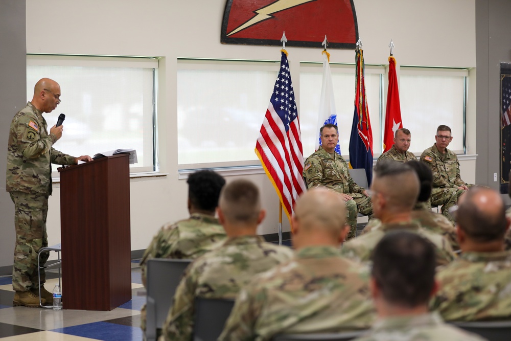 A military ceremony or briefing is taking place indoors, with a soldier speaking at a podium while three senior personnel are seated on a dais behind him. An audience of uniformed soldiers sits facing the speaker. The setting appears to be an official military function room decorated with flags and a unit insignia on the wall.