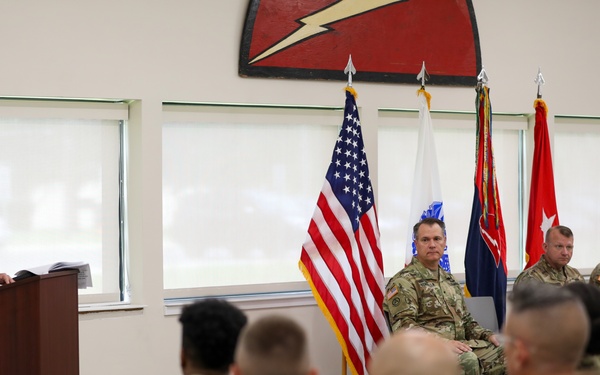 A military ceremony or briefing is taking place indoors, with a soldier speaking at a podium while three senior personnel are seated on a dais behind him. An audience of uniformed soldiers sits facing the speaker. The setting appears to be an official military function room decorated with flags and a unit insignia on the wall.