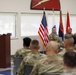 A military ceremony or briefing is taking place indoors, with a soldier speaking at a podium while three senior personnel are seated on a dais behind him. An audience of uniformed soldiers sits facing the speaker. The setting appears to be an official military function room decorated with flags and a unit insignia on the wall.