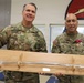 Two U.S. Army soldiers in MultiCam/OCP camouflage uniforms are posing together while holding a ceremonial saber mounted on a wooden display plaque. The soldier on the left is identified by name tape as 'SCARCELLA' and wears a Brigadier General star insignia, while the soldier on the right is identified as 'FARABAUGH' and wears a Colonel eagle insignia along with multiple medals including what appears to be a Meritorious Service Medal. This appears to be a military retirement or award ceremony presentation.