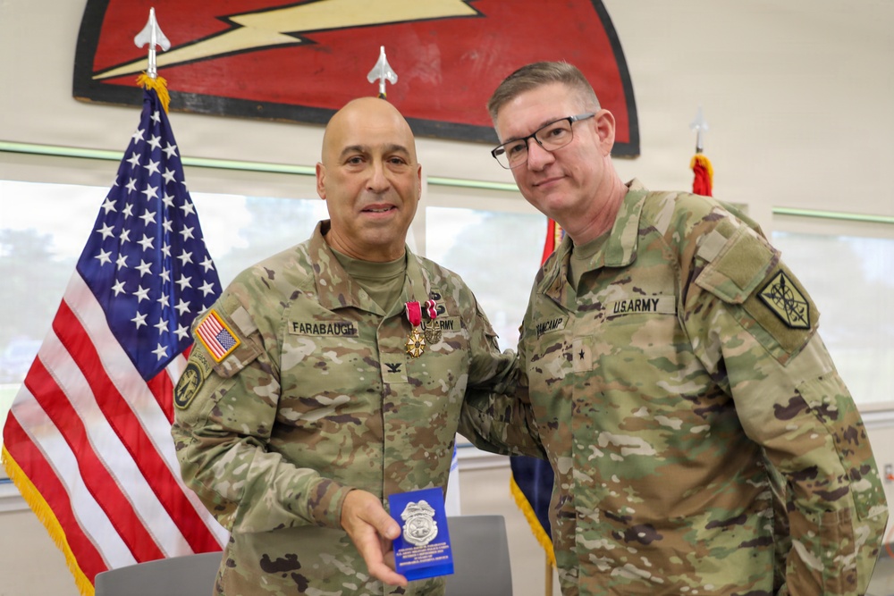 Two U.S. Army soldiers pose together in a formal military ceremony setting. The soldier on the left, identified by name tape as 'FARABAUGH,' is wearing a medal (appears to be a Meritorious Service Medal or similar decoration) and holding a small blue case, suggesting a recent award presentation. The soldier on the right, identified as 'VANCAMP,' displays a two-star general rank insignia on his collar.