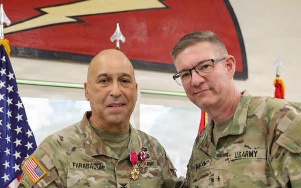 Two U.S. Army soldiers pose together in a formal military ceremony setting. The soldier on the left, identified by name tape as 'FARABAUGH,' is wearing a medal (appears to be a Meritorious Service Medal or similar decoration) and holding a small blue case, suggesting a recent award presentation. The soldier on the right, identified as 'VANCAMP,' displays a two-star general rank insignia on his collar.