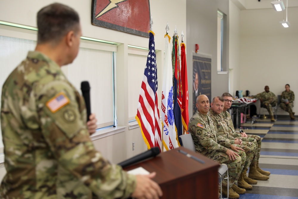 A military ceremony or briefing is taking place indoors, with a uniformed soldier speaking at a podium while holding a microphone. Several other soldiers in Army Combat Uniforms (ACU/OCP pattern) are seated in chairs along the wall, listening attentively. Multiple flags, including the U.S. flag and military unit colors, are displayed in the background.