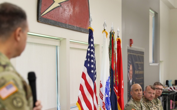 A military ceremony or briefing is taking place indoors, with a uniformed soldier speaking at a podium while holding a microphone. Several other soldiers in Army Combat Uniforms (ACU/OCP pattern) are seated in chairs along the wall, listening attentively. Multiple flags, including the U.S. flag and military unit colors, are displayed in the background.