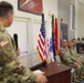 A military ceremony or briefing is taking place indoors, with a uniformed soldier speaking at a podium while holding a microphone. Several other soldiers in Army Combat Uniforms (ACU/OCP pattern) are seated in chairs along the wall, listening attentively. Multiple flags, including the U.S. flag and military unit colors, are displayed in the background.