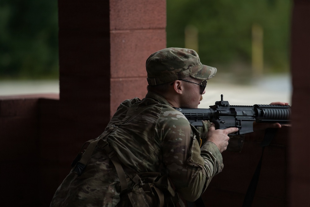 A soldier in MultiCam/OCP camouflage uniform is positioned behind a brick column, aiming a rifle in a tactical stance. The individual is wearing a patrol cap, eye protection, and carrying a backpack. The setting appears to be a training environment or military installation based on the structured surroundings.
