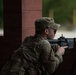 A soldier in MultiCam/OCP camouflage uniform is positioned behind a brick column, aiming a rifle in a tactical stance. The individual is wearing a patrol cap, eye protection, and carrying a backpack. The setting appears to be a training environment or military installation based on the structured surroundings.