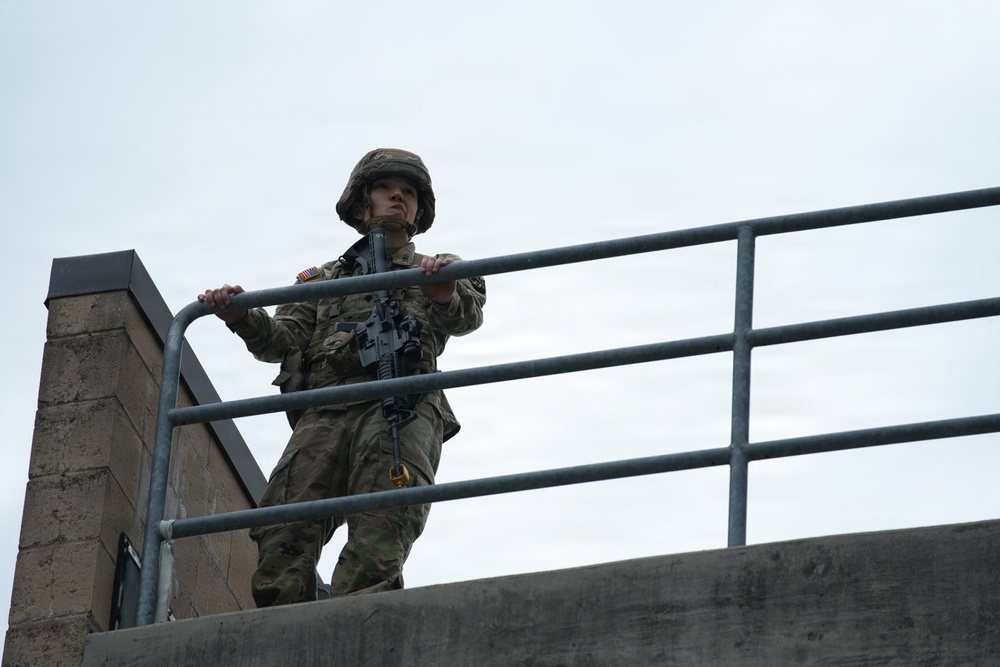 A U.S. Army soldier in full combat uniform stands on an elevated concrete structure, gripping a metal railing while scanning the surroundings. The soldier is equipped with a rifle, combat helmet, and OCP (Operational Camouflage Pattern) uniform with a visible American flag patch on the shoulder. The low-angle photograph emphasizes the soldier's elevated position against an overcast sky.