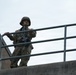 A U.S. Army soldier in full combat uniform stands on an elevated concrete structure, gripping a metal railing while scanning the surroundings. The soldier is equipped with a rifle, combat helmet, and OCP (Operational Camouflage Pattern) uniform with a visible American flag patch on the shoulder. The low-angle photograph emphasizes the soldier's elevated position against an overcast sky.