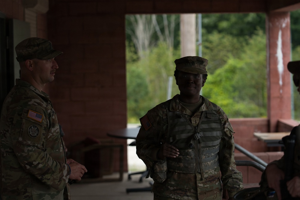 Two U.S. Army soldiers in MultiCam (OCP) uniforms are standing under a covered structure, appearing to be in conversation. Both wear patrol caps and have U.S. flag patches visible on their left shoulders. The setting appears to be an outdoor training or administrative area with trees visible in the background.