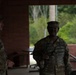 Two U.S. Army soldiers in MultiCam (OCP) uniforms are standing under a covered structure, appearing to be in conversation. Both wear patrol caps and have U.S. flag patches visible on their left shoulders. The setting appears to be an outdoor training or administrative area with trees visible in the background.