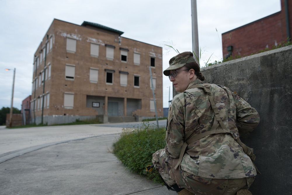 A soldier in full MultiCam/OCP camouflage uniform is crouching against a concrete barrier, appearing to take cover or observe surroundings. The setting appears to be an urban training environment featuring a large, abandoned multi-story brick building with boarded or missing windows. The scene suggests a military training exercise in a mock urban environment.