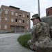 A soldier in full MultiCam/OCP camouflage uniform is crouching against a concrete barrier, appearing to take cover or observe surroundings. The setting appears to be an urban training environment featuring a large, abandoned multi-story brick building with boarded or missing windows. The scene suggests a military training exercise in a mock urban environment.