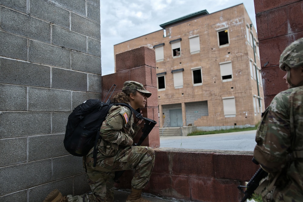Two soldiers in U.S. Army camouflage uniforms are conducting what appears to be a military training exercise in an urban environment. One soldier is crouched in a low position against a cinder block wall, peering around a corner toward a multi-story brick building, while a second soldier stands nearby. The setting resembles a Military Operations on Urban Terrain (MOUT) training facility.