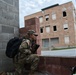 Two soldiers in U.S. Army camouflage uniforms are conducting what appears to be a military training exercise in an urban environment. One soldier is crouched in a low position against a cinder block wall, peering around a corner toward a multi-story brick building, while a second soldier stands nearby. The setting resembles a Military Operations on Urban Terrain (MOUT) training facility.