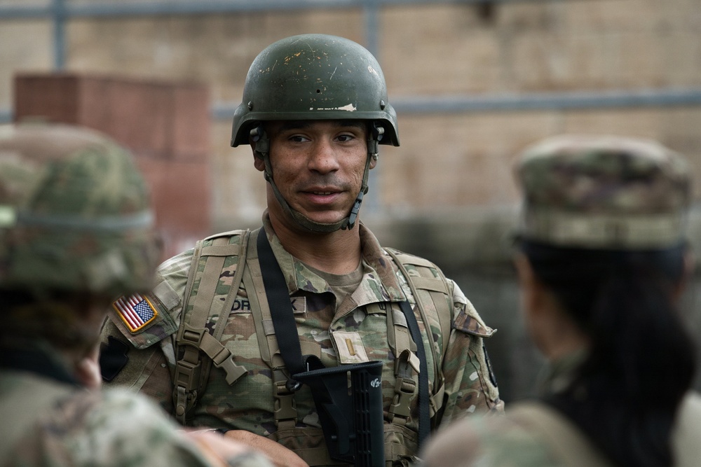 A U.S. Army soldier wearing OCP (Operational Camouflage Pattern) uniform and an olive drab helmet is engaged in conversation with other soldiers. The central figure displays a U.S. flag patch on the left shoulder and appears to hold a rank insignia on the chest. The setting appears to be an outdoor military training or operational environment.