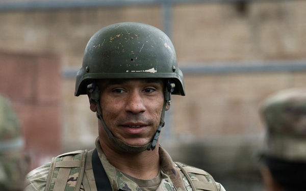 A U.S. Army soldier wearing OCP (Operational Camouflage Pattern) uniform and an olive drab helmet is engaged in conversation with other soldiers. The central figure displays a U.S. flag patch on the left shoulder and appears to hold a rank insignia on the chest. The setting appears to be an outdoor military training or operational environment.