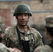 A U.S. Army soldier wearing OCP (Operational Camouflage Pattern) uniform and an olive drab helmet is engaged in conversation with other soldiers. The central figure displays a U.S. flag patch on the left shoulder and appears to hold a rank insignia on the chest. The setting appears to be an outdoor military training or operational environment.