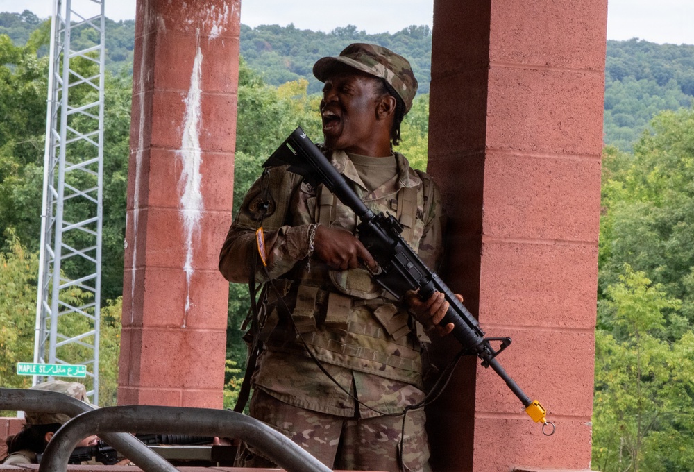 A soldier in US Army MultiCam (OCP) camouflage uniform is holding a rifle with a yellow training/blank firing attachment on the muzzle, indicating a training exercise. The individual is smiling/laughing and positioned between red brick or concrete columns in what appears to be a military training facility. A bilingual street sign reading 'Maple St' in English and Arabic is visible in the background, suggesting a mock urban warfare training environment.