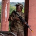 A soldier in US Army MultiCam (OCP) camouflage uniform is holding a rifle with a yellow training/blank firing attachment on the muzzle, indicating a training exercise. The individual is smiling/laughing and positioned between red brick or concrete columns in what appears to be a military training facility. A bilingual street sign reading 'Maple St' in English and Arabic is visible in the background, suggesting a mock urban warfare training environment.