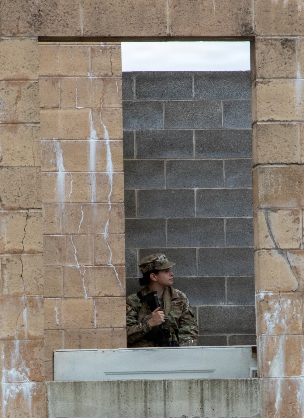A U.S. Army soldier in full camouflage uniform and patrol cap is visible through a rectangular window or doorway opening in a weathered stone wall. The soldier is holding a rifle and appears to be in a tactical or training posture, looking to one side. The setting appears to be a military training facility with mixed construction materials including sandstone and concrete block walls.