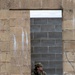 A U.S. Army soldier in full camouflage uniform and patrol cap is visible through a rectangular window or doorway opening in a weathered stone wall. The soldier is holding a rifle and appears to be in a tactical or training posture, looking to one side. The setting appears to be a military training facility with mixed construction materials including sandstone and concrete block walls.