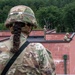 Three soldiers in OCP (Operational Camouflage Pattern) uniforms are conducting what appears to be a military training exercise at a mock urban structure. The foreground soldier, identifiable as female by her braided hair, observes two other soldiers positioned on the rooftop of a red brick building. One rooftop soldier is aiming a rifle while the other stands in an overwatch position.