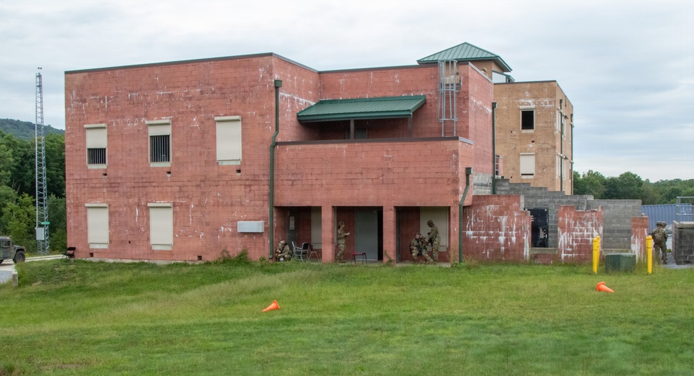 A military training exercise is taking place at what appears to be a mock urban operations facility or MOUT (Military Operations on Urban Terrain) site. Several soldiers in combat gear are positioned around the entrance of a weathered two-story red brick building, conducting a building clearance or entry drill. Orange traffic cones are placed on the grass field in front of the structure, likely marking boundaries or safety zones.