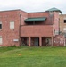 A military training exercise is taking place at what appears to be a mock urban operations facility or MOUT (Military Operations on Urban Terrain) site. Several soldiers in combat gear are positioned around the entrance of a weathered two-story red brick building, conducting a building clearance or entry drill. Orange traffic cones are placed on the grass field in front of the structure, likely marking boundaries or safety zones.