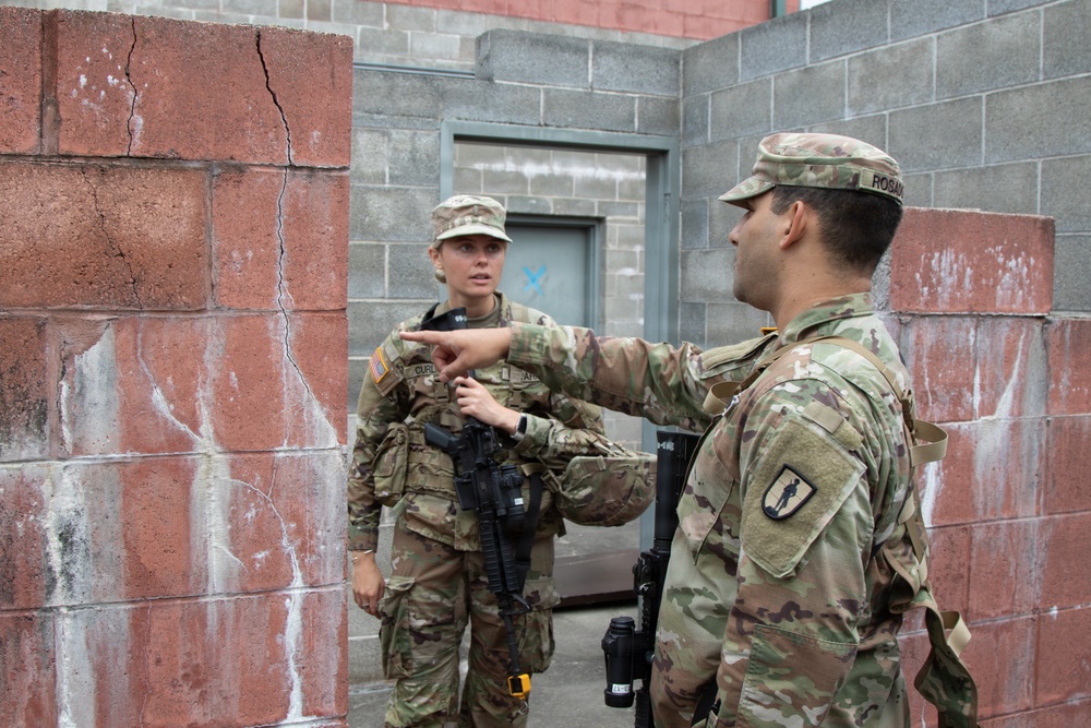 Two U.S. Army soldiers in Operational Camouflage Pattern (OCP) uniforms are engaged in what appears to be a training exercise or briefing in an urban operations environment. A male soldier with the name tape 'ROSAS' is pointing and providing instruction to a female soldier, both carrying rifles. The setting appears to be a military training facility with concrete block walls simulating an urban combat environment.