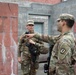 Two U.S. Army soldiers in Operational Camouflage Pattern (OCP) uniforms are engaged in what appears to be a training exercise or briefing in an urban operations environment. A male soldier with the name tape 'ROSAS' is pointing and providing instruction to a female soldier, both carrying rifles. The setting appears to be a military training facility with concrete block walls simulating an urban combat environment.